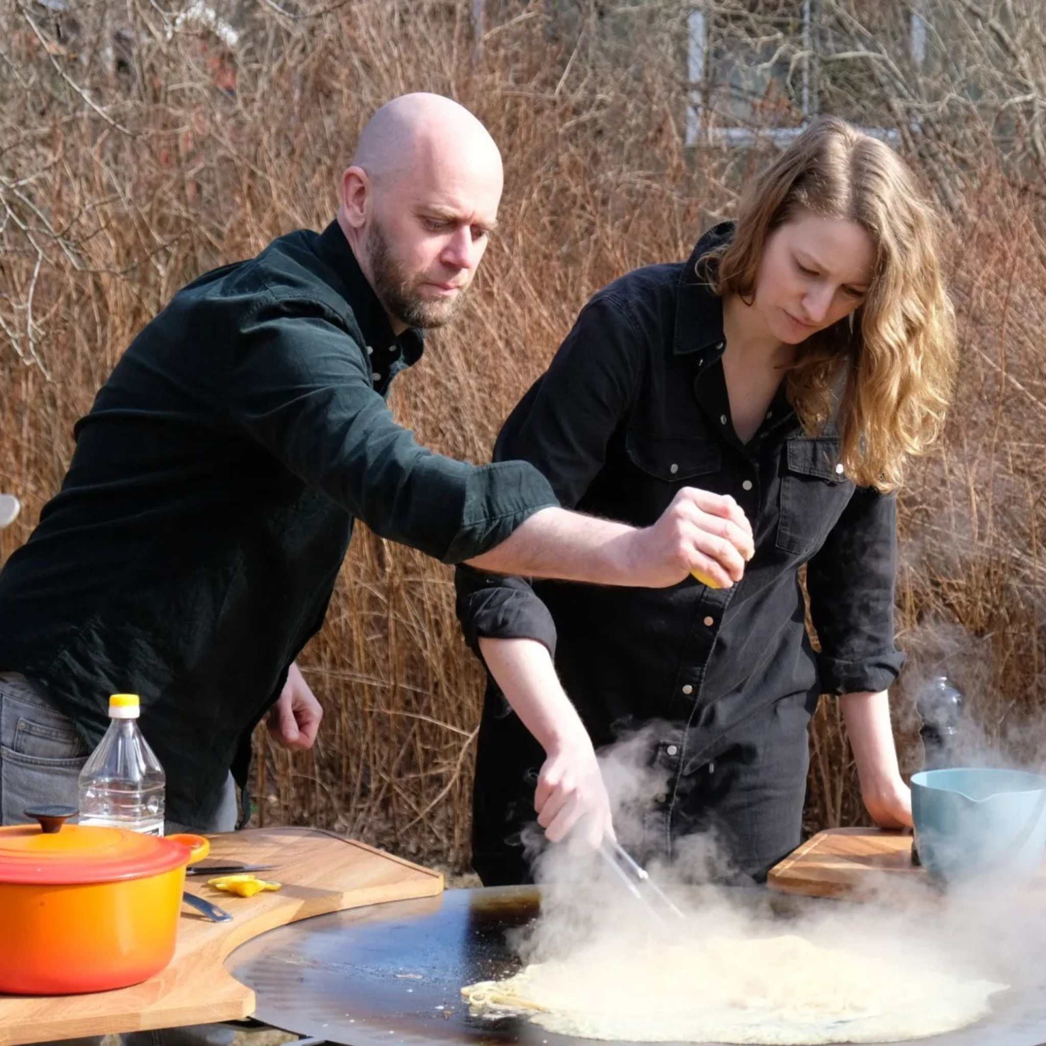 Kokbok för Stekhäll, kokbok med recept för matlagning på stekhäll, där två personer lagar mat utomhus.