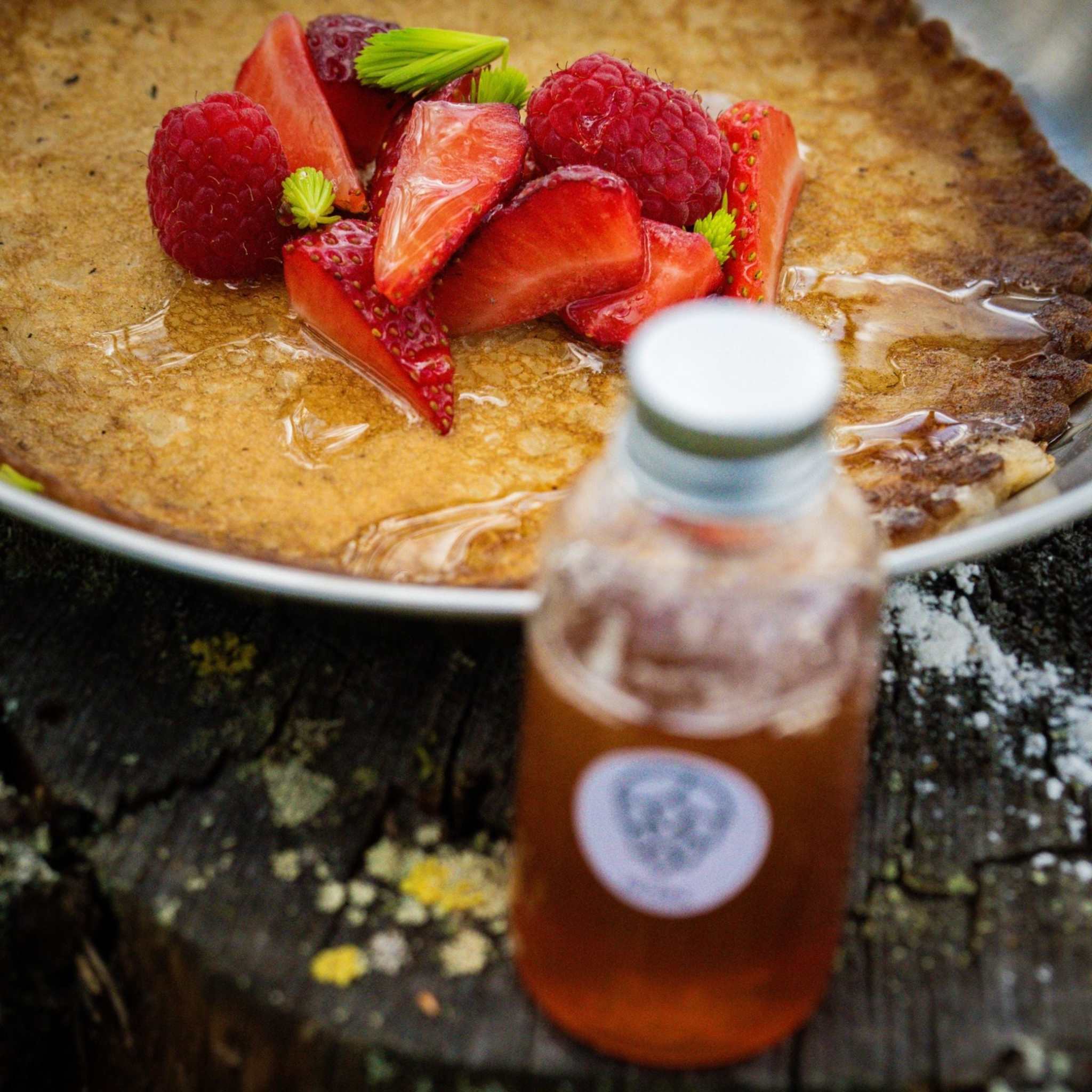 Pannkaku med Granskottsirap med gyllenbrun pannkaka, röda bär och en flaska bärnstensfärgad sirap.