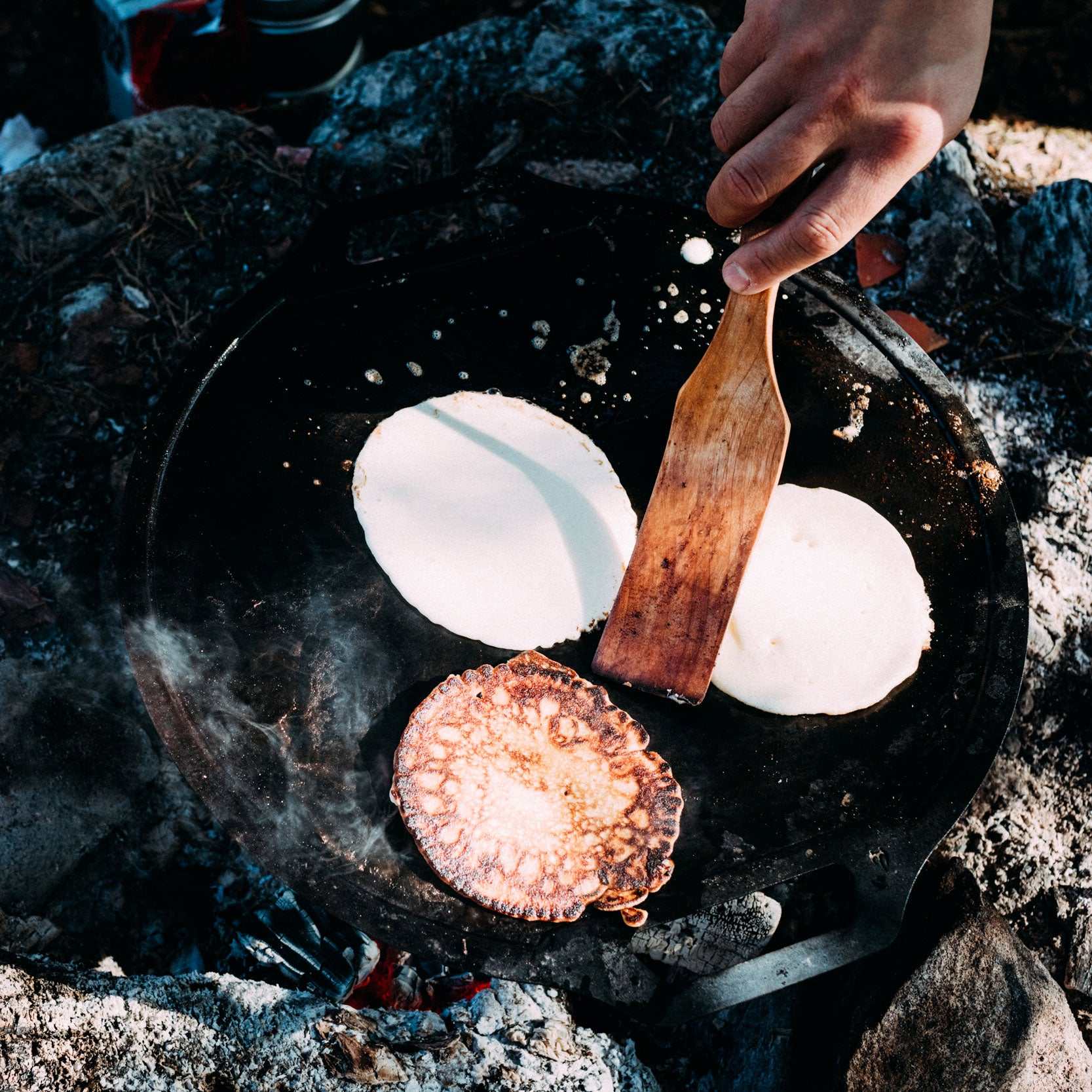 Pannkakor steks på svart stekhäll med träspatel; Pannkaku med Granskottsirap för matlagning utomhus.