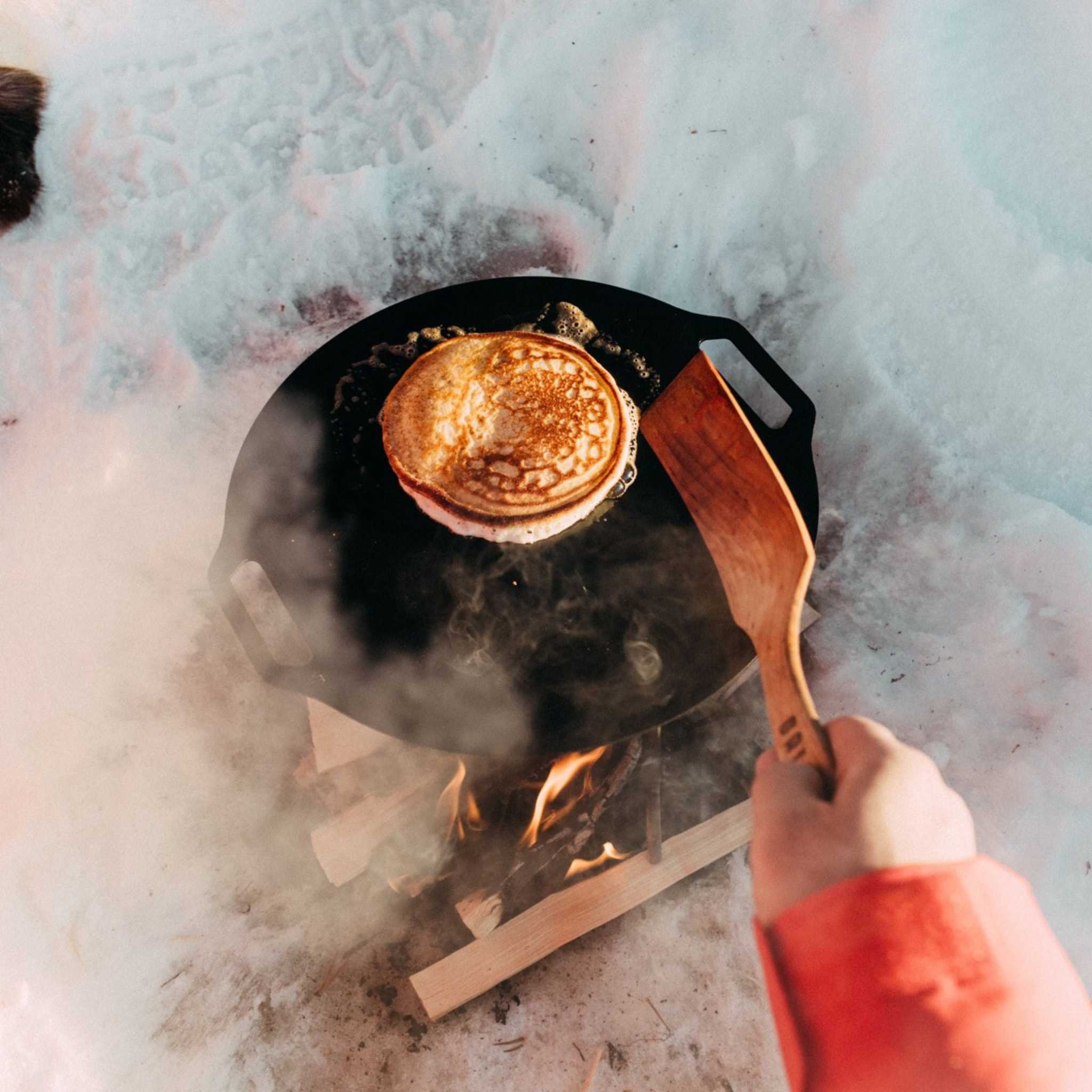 Pannkaku med Lingonsirap med gyllenbrun pannkaka i svart stekhäll, vänd med en träspatel över öppen eld.