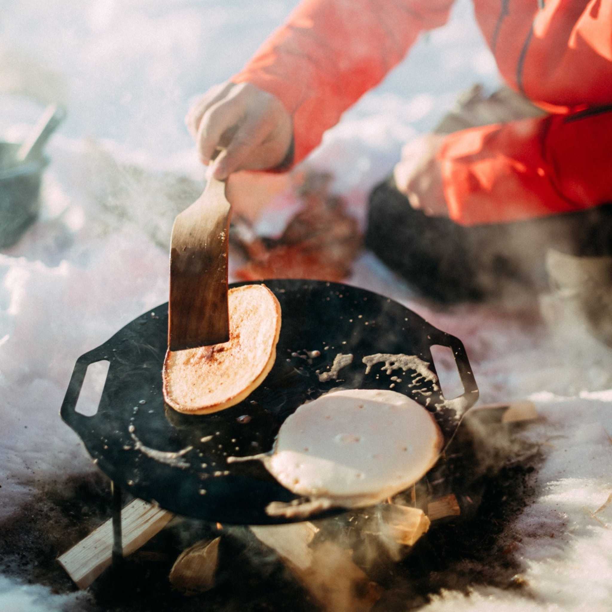 Pannkakor på svart stekhäll, gyllenbruna och ljusa, vänds med stekspade; Pannkaku med Lingonsirap passar ute i naturen.