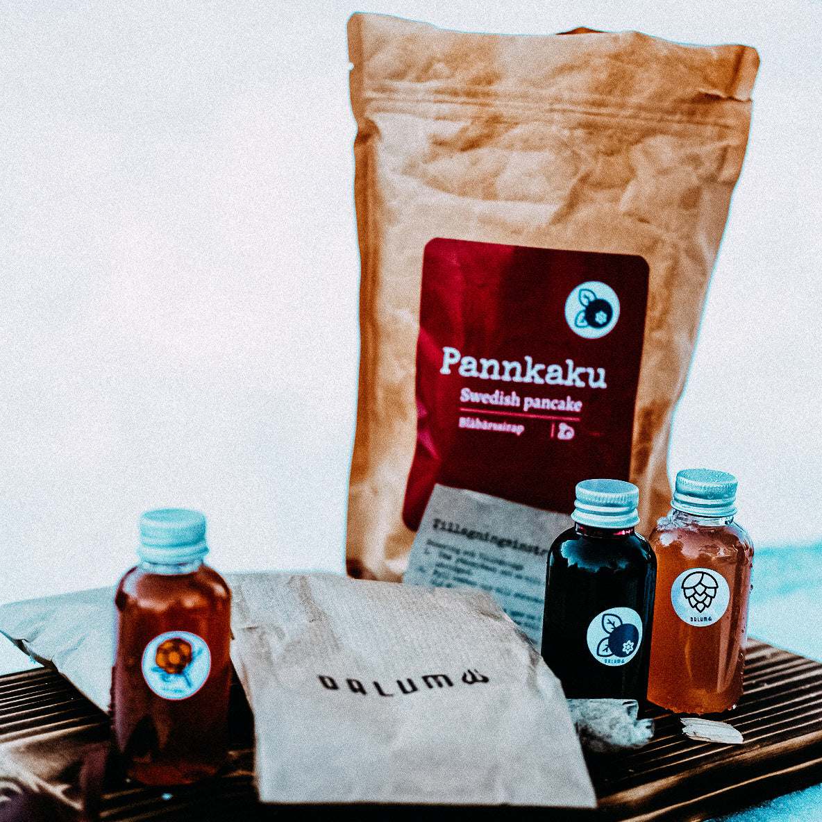 Pannkaku med Lingonsirap, brun papperspåse med röd etikett och små sirapsflaskor i genomskinlig plast.
