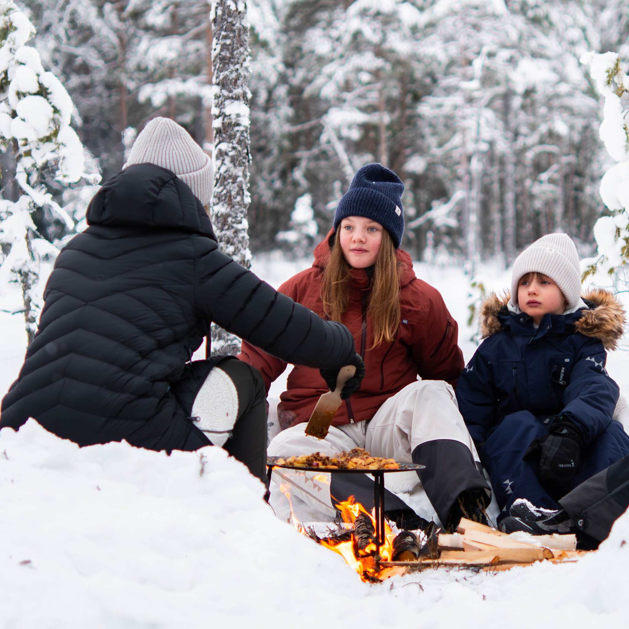 Dalum stekhäll i mörkt stål med tre ben, används över öppen eld för matlagning utomhus i vintermiljö.