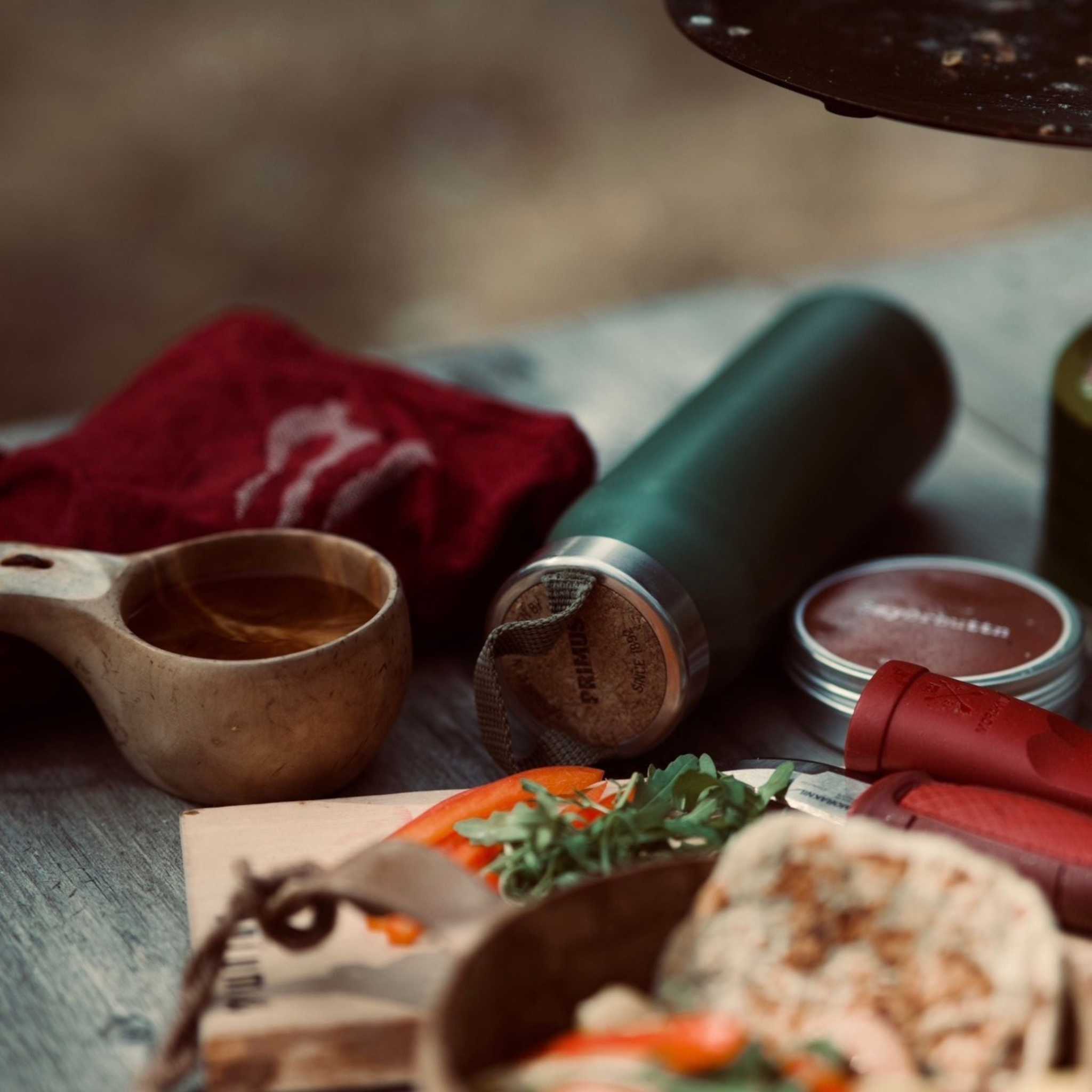 Linnehandduk outdoor, vinröd handduk i linnetyg med ljus logotyp, vikt bredvid en träkåsa, termos och matvaror.